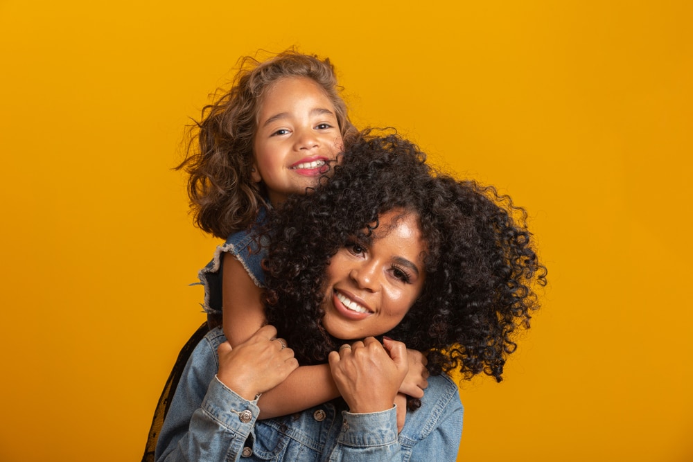 A mother wearing a jean jacket holds her daughter on her shoulders and they smile for the camera in front of an orange background