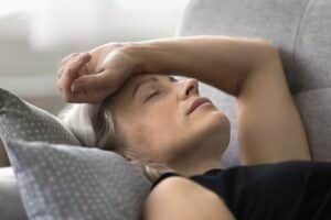 Senior woman lying on a sofa with her arm on her head, appearing fatigued and dizzy, illustrating symptoms of menopause or low blood pressure, such as headache and daytime exhaustion.